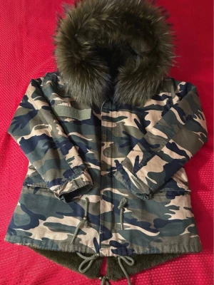 Camouflage  vinterjacka med naturlig päls  - Vinterjacka i camouflage med stor huva och fluffig naturlig päls . Insidan är fodrad med mjuk grön pälsimitation. Jackan har snörning i midjan och nederkant, dragkedja och knappar framtill. Perfekt för kalla dagar och riktigt snygg streetstil.