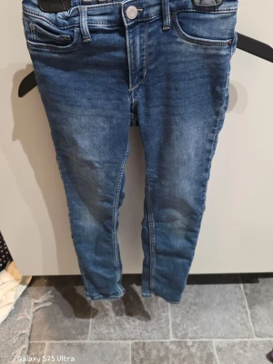Blå skinny jeans i denim, strl 134 - Säljer ett par blå skinny fit jeans från Denim & Denim i storlek 134. Jeansen har klassisk femficksdesign, knapp och dragkedja fram samt justerbar midja med resår. Materialet är en mix av bomull, polyester och elastan för skön stretch. Perfekt passform för dig som gillar smala jeans.
