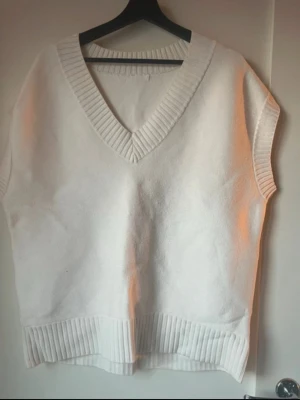 Vit stickad väst med v-ringning - Säljer en stilren vit stickad väst med bred ribbad v-ringning och ribbade kanter. Västen har en loose passform och är perfekt att bära över en skjorta eller t-shirt för en trendig lager-på-lager-look.