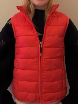 Röd dunväst från CRW - Säljer en röd dunväst från CRW i storlek 146/152. Västen har quiltad design, hög krage och dragkedja framtill. Perfekt att slänga över en hoodie för extra värme. Två fickor med dragkedja på sidorna. Enkel och clean look.
