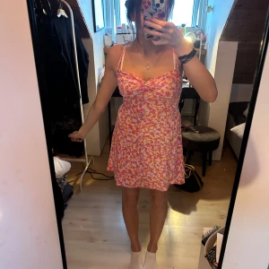 Färglad blommig kort klänning med smala axelband - Säljer en söt kort klänning med blommigt mönster i rosa, orange och vitt. Klänningen har smala axelband och en lätt v-ringad överdel med twistad detalj framtill. Perfekt för varma dagar och har en luftig passform.