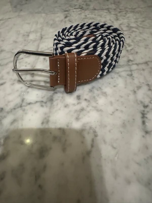 Blåvit flätad bälte med skinn-detaljer  - Snyggt flätat bälte i marinblått och vitt med bruna skinn-detaljer vid spännet och änden. Bältet har ett klassiskt silverfärgat metallspänne och kontrastsömmar som ger en stilren look. Perfekt för att lyfta din outfit med en cool touch.