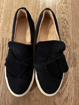 Svarta mocka loafers med rosett - Säljer ett par svarta loafers från Billibi i mjuk mocka. Skorna har en stor dekorativ rosett framtill och vit platt sula. Perfekta för dig som vill ha en stilren men ändå unik look.