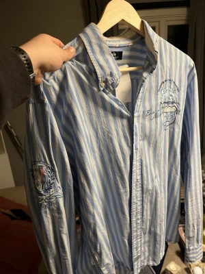 Blåvit randig skjorta La Martina XL - Säljer en blå och vit randig skjorta från La Martina i storlek XL. Skjortan har broderade detaljer på bröstet och ärmen med polo-inspirerade emblem och text. Klassisk krage och knappar framtill. Perfekt för dig som gillar sportig och preppy stil.