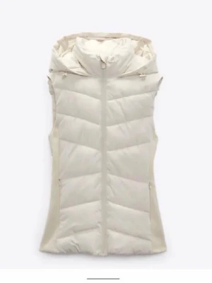 Vit dunväst med huva från Zara - Säljer en snygg vit dunväst från Zara i storlek XS. Den är använd men ser ändå bra ut. Västen har en stor, mysig huva och dragkedja framtill. Quiltad design med breda paneler och mjuka sidor i beige. Perfekt att slänga över en hoodie när det är kyligt ute.