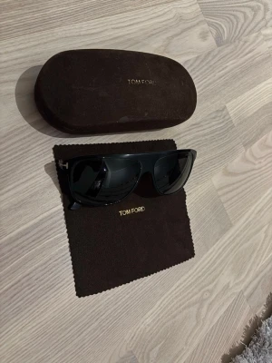 Svart Tom Ford solglasögon - Säljer ett par snygga svarta solglasögon från Tom Ford med klassisk fyrkantig form och diskret guldfärgad detalj vid skalmen. Glasögonen har mörka linser och kommer med originalfodral och putsduk. Perfekt accessoar för en cool look.