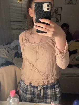 Beige blus med volangdetaljer - Säljer en söt beige blus med volangdetaljer längs halsringningen, diagonalt över framsidan och vid fållen. Blusen har lång ärm och är tillverkad i ett lätt, skrynkligt tyg som ger en avslappnad känsla. Perfekt att styla med jeans eller kjol. Står inte vart den kommer ifrån eller vilken storlek, men tröjan är ganska tajt så gissar på XS