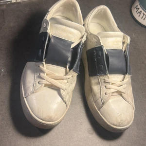 Valentino Garavani sneakers vit/blå - Snygga sneakers från Valentino Garavani i vitt skinn med bred mörkblå rem över snörningen och coola nitar på hälen. Klassisk rund tå och platt sula, perfekt för dig som gillar exklusiv streetstil. Skorna har vita snören och en chunky siluett. Sulan är byt.