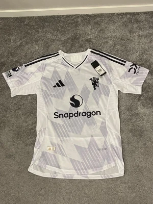 Manchester United Tröja - Säljer en vit Manchester United fotbollströja med Mbeumo 19 på ryggen. Tröjan har svarta och lila detaljer, randigt mönster och meshpaneler för extra ventilation. Adidas-logga och Snapdragon sponsortryck på framsidan. Slim fit och autentisk licensierad produkt. Helt ny tröja, testad en gång