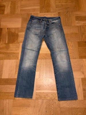 Levi's 501 blå jeans W28 L32 - Säljer ett par klassiska Levi's 501 jeans i blå tvätt med raka ben och normal passform. Jeansen har fem fickor, knappgylf och den ikoniska läderpatchen bak i midjan. Perfekt för dig som gillar tidlös stil och äkta denim.