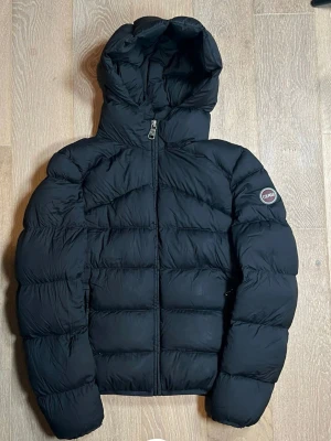 Svart pufferjacka från Colmar - Säljer en svart pufferjacka från Colmar med huva och dragkedja framtill. Jackan har ett stilrent, quiltat mönster och en rund logotyp på ärmen. Perfekt för kalla dagar och har en snygg, modern look som passar till streetwear.