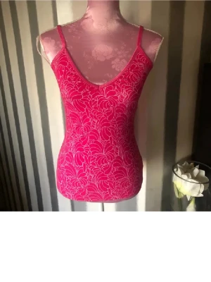 Rosa blommig linnetopp - Snygg rosa linnetopp med tunna axelband och blommigt mönster i vitt. Toppen har en v-ringad hals och sitter figurnära. Perfekt för varma dagar och enkel att styla med jeans eller kjol. reglerbara axelband och inbyggd bh. Storlek M på lappen men mindre i storleken . Ny med prislappen på 