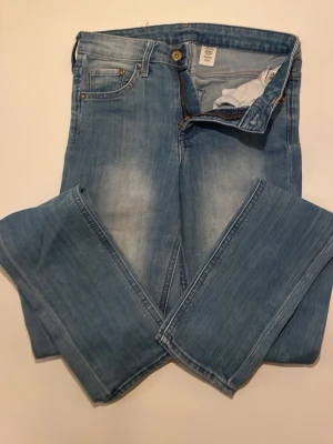 Ljusblå raka jeans med bältesdetalj - Säljer ett par ljusblå jeans med raka ben och snygg tvätt. Jeansen har klassiska fem fickor, dragkedja och knapp i guldton samt en unik bältesdetalj i midjan. Materialet är jeans och passformen är normal. Perfekta till en avslappnad look. 27/32 skinny med stretch 