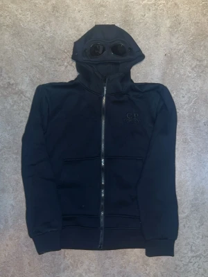 Svart C.P Company hoodie - Svart C.P. Company hoodie med dragkedja Stilren och bekväm hoodie från C.P. Company i klassisk svart färg. Tillverkad i mjuk och högkvalitativ bomullsblandning för skön komfort och bra passform. Hoodien har hel dragkedja framtill, justerbar huva med snörning samt ribbade muddar vid ärmslut och nederkant. Ikonisk C.P. Company-design med fokus på kvalitet och streetwear-stil. Perfekt för både vardag och avslappnade outfits.