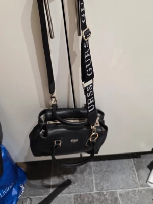 Svart handväska från Guess - Snygg svart handväska från Guess med stilrena guldfärgade detaljer och logotyp framtill. Väskan har både handtag och en bred axelrem med tydlig Guess-text. Insidan är ljus med praktisk dragkedja och flera fack. Perfekt för dig som gillar klassisk design med en modern touch.