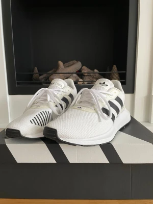 Adidas sneakers vita med svarta ränder - Fräscha Adidas sneakers i vitt med klassiska svarta ränder på sidorna och mesh-ovandel. Skorna har snörning, rund tå och platt sula. Märket syns tydligt på plösen och hälen. Snygga detaljer med svarta linjer på ena skon. 