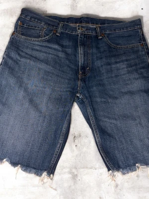 Levi's jeansshorts med rå kant - Säljer ett par klassiska jeansshorts från Levi's i mörkblå denim. De har fem fickor, normal passform och rå, fransig kant nedtill för en chill vibe. Perfekta för sommaren och har den ikoniska röda Levi's-taggen på bakfickan.