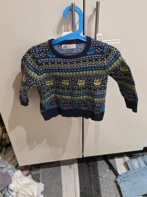 Mönstrad blå stickad tröja H&M - Säljer en cool stickad tröja från H&M i storlek 86/92. Tröjan är mörkblå med färgglada mönster i gult, blått, orange och vitt. Rund hals och långa ärmar, tillverkad i 100% bomull. Perfekt för dig som gillar färg och mönster!