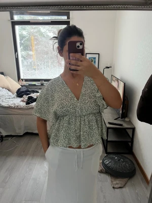 Blommig v-ringad blus med volang - Säljer en söt blommig blus i vitt och grönt med v-ringning och volangdetaljer. Blusen har korta ärmar och en luftig, croppad passform som ger en avslappnad vibe. Perfekt för varmare dagar och enkel att styla med både kjol och jeans.