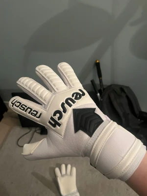 Vita målvaktshandskar från Reusch - Säljer ett par vita målvaktshandskar från Reusch med svart logga och detaljer. Handskarna har vadderade fingrar och bred kardborre vid handleden för extra stöd. Insidan är slät och greppvänlig, perfekt för fotbollsmålvakter som vill ha bra kontroll och komfort. Helt oanvända och plasten. Sitter fortfarande kvar på latexen. Storlek 10 