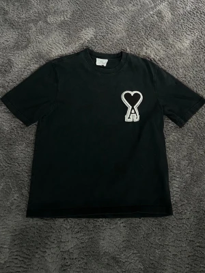 Svart t-shirt från Ami med hjärtdetalj - Svart t-shirt från Ami med klassisk passform och korta ärmar. På bröstet sitter en stor broderad patch med ett hjärta och bokstaven A i vitt och svart. Materialet är mjuk bomull och halsen är rundad. Perfekt för dig som gillar stilrena plagg med en twist.