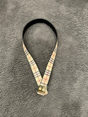 Rutig Burberry Belte - Säljer ett snyggt vändbart skärp med klassiskt rutigt mönster i beige, svart och rött på ena sidan och helsvart med präglat mönster på andra sidan. Skärpet har ett guldfärgat B-spänne! Priset kan diskuteras! 