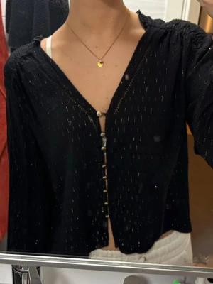 Svart glittrig blus - Elegant svart blus med v-ringning och dekorativa knappar framtill. Långärmad modell i ett mjukt, följsamt material med diskret struktur som ger en exklusiv känsla. Passar lika bra till vardag som till fest – enkel att styla med jeans, kjol eller kostymbyxor.