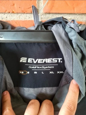 Svart anorakjacka från Everest Xs S - Snygg svart anorakjacka från Everest med huva och snörning. Jackan har en stor ficka framtill med dragkedja och tryckknapp, samt justerbara ärmslut. Perfekt för blåsiga dagar och outdoor-aktiviteter. GoldFlexSystem för extra komfort.