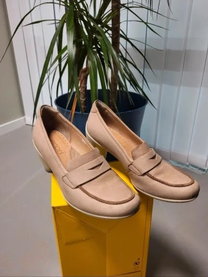 Beige loafers med klack från Ecco 37 - Snygga beige loafers från Ecco med rund tå och låg klack. Skorna är tillverkade i mjukt skinn och har klassisk loafer-design med dekorativ rem över foten. Perfekta för dig som gillar stilrena och bekväma skor med lite höjd.