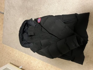 Svart dunjacka från Canada Goose - Svart dunjacka från Canada Goose med klassisk röd och vit logotyp på ärmen. Jackan har huva, dragkedja och knappar framtill. Fylld med dun och fjädrar för extra värme, perfekt för kalla dagar. Snygg och stilren design som funkar till alla dina vinteroutfits. Med päls tillkommer såklart