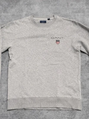 Grå tröja från GANT - Säljer en stilren grå sweatshirt från GANT med broderad logga och sköld på bröstet. Tröjan har rund hals, ribbade muddar och är tillverkad i mjuk bomullsmix. Perfekt för en clean och avslappnad look. Storlek 146-152.