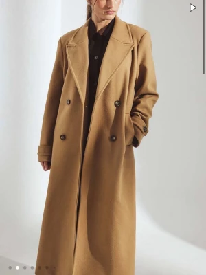 Kamelbrun lång kappa med knappar - Snygg lång kappa i kamelbrun färg med dubbelknäppning och breda slag. Klassisk design med raka linjer och fickor på sidorna. Perfekt för dig som vill ha en stilren och tidlös look. Materialet känns mjukt och värmande, passar bra till höst och vinter. Helt ny och nypris 1400