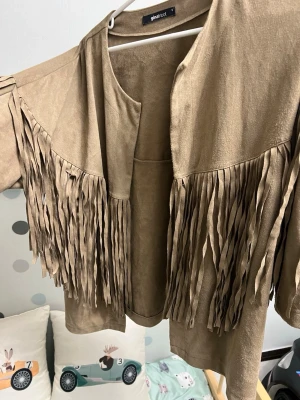 Beige mocka overshirt med fransar - Säljer en beige overshirt från Gina Tricot i mjuk mocka-look med långa fransar längs axlar och rygg. Jackan har öppen front och loose passform, perfekt för dig som vill sticka ut med cool western-vibe. Passar till jeans och boots för en avslappnad stil.