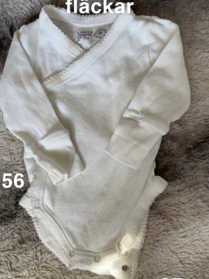 Vit omlottbody från Lindex - Söt vit omlottbody från Lindex med små diskreta hålmönster och spetskant. Lång ärm och tryckknappar i grenen för enkel påklädning. Perfekt till de allra minsta.🌸Fläckar finns. Storlek 56