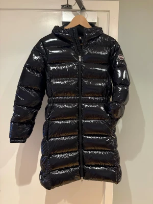 Svart glansig dunjacka från Colmar (med ett litet hål) - Säljer en svart, glansig lång dunjacka från Colmar med huva och tvåvägsdragkedja. Jackan har puffig design, emblem på ärmen och är perfekt för kalla dagar. Tillverkad i syntetmaterial med dunfyllning för extra värme.