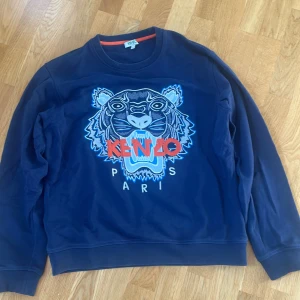 Mörkblå Kenzo sweatshirt med tiger - Snygg mörkblå sweatshirt från Kenzo med stort broderat tigerhuvud och logga i rött och blått på bröstet. Tröjan har rund hals, långa ärmar och ribbade muddar. Perfekt för dig som vill sticka ut lite extra! Populär, snygg och fullt äkta.