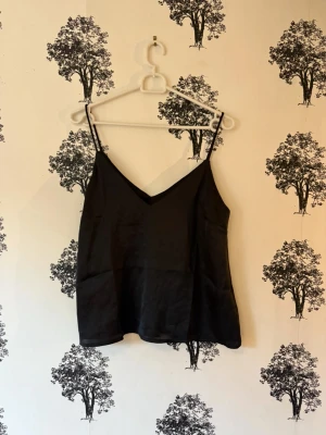 Svart linnetopp  - Säljer en stilren svart linnetopp från The Black Label Collection från Lager 157  i storlek S. Toppen har tunna axelband och en v-ringad hals, vilket ger en enkel och snygg look. Materialet är mjukt och har en lätt glansig finish som känns lyxig.