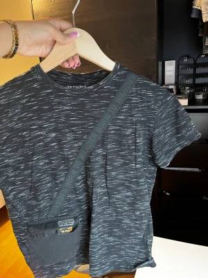 Svart t-shirt med ficka från Pantaloons - Cool svart t-shirt från Pantaloons med vita streck som ger en unik look. T-shirten har en praktisk ficka framtill med textdetalj och ett band med trycket 'Born to Wander'. Perfekt för dig som gillar streetstyle och vill sticka ut lite extra.