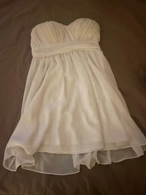 Vit axelbandslös kortklänning Model Behaviour - Säljer en vit axelbandslös kortklänning från Model Behaviour i storlek 34. Klänningen har ett draperat liv och ett flowigt, lager-på-lager kjolparti. Perfekt för dig som gillar romantisk och stilren design. Materialet känns lätt och luftigt.