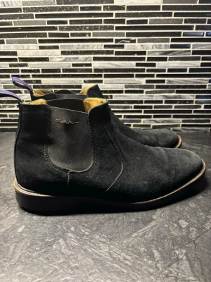 Svarta chelsea boots i mocka från Gant - Säljer ett par svarta chelsea boots från Gant i mjuk mocka. Skorna har elastiska paneler på sidorna, rund tå och robust gummisula med greppmönster. Insidan är fodrad i ljust skinn och de har en snygg blå-röd hälflik för enkel påtagning.