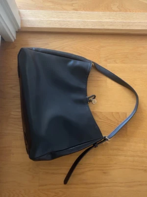 Svart axelväska i skinn - Stilren svart axelväska med mjuk, slät yta i skinn och bred axelrem. Väskan är från zalando och har en enkel design med guld detalj. Perfekt storlek för att få plats med det viktigaste. 