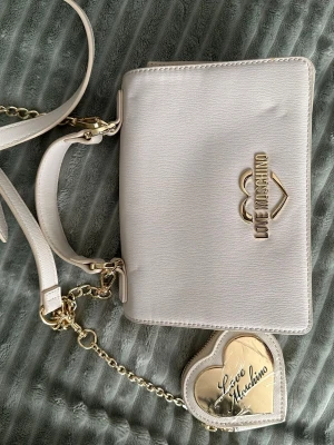 Vit axelväska från Love Moschino - Säljer en stilren vit axelväska från Love Moschino med guldfärgade detaljer och hjärtformad nyckelring. Väskan har en slät struktur, magnetlås och justerbar axelrem i skinn. Perfekt för dig som gillar snygga accessoarer med exklusiv känsla.