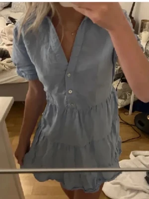 Ljusblå volangklänning från Zara - Säljer en ljusblå kortklänning från Zara med volanger och v-ringning. Klänningen har korta ärmar, knappar framtill och en avslappnad passform. Perfekt för varma dagar och har en mjuk bomullskänsla.