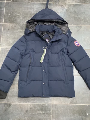 Mörkblå dunjacka från Canada Goose - Säljer en mörkblå dunjacka från Canada Goose med avtagbar huva och klassisk patch på ärmen. Jackan har dragkedja och knappar framtill, långa ärmar och är fodrad med svart material på insidan. Perfekt för kalla dagar och har en clean, stilren look.
