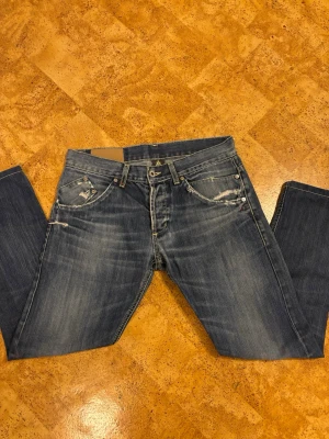 Dondup jeans med slitningar - Säljer ett par blå dondup jeans med snygga slitningar och detaljerade sömmar. Jeansen har klassisk femficksdesign, raka ben och coola slitna partier både fram och bak. Märkeslogga bak i midjan och kontrastsömmar för extra stil.