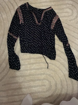 Blommig blus från BikBok XS - Mörkblå blus från BikBok i storlek XS med små blommiga mönster i vitt, rosa och blått. Blusen har långa ärmar, v-ringad hals och dekorativa detaljer i rosa vid axlar och hals. Knytband i midjan för extra passform.