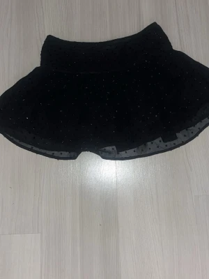 Svart kortkjol med glitterprickar - Supersöt svart kortkjol med glittriga prickar och volang från Gina Young! ✨Kjolen har ett brett, veckat midjeband som ger en snygg siluett. Tunt, lätt material som känns luftigt. Perfekt för dig som vill sticka ut lite extra. 🪩Storlek 146/152, använd endast en gång så jättefint skick! ⭐️