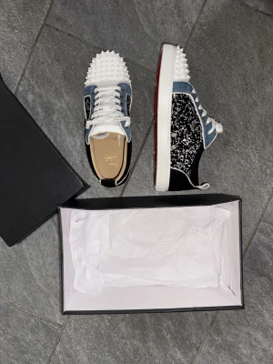 Christian Louboutin sneakers med nitar - Säljer ett par unika sneakers från Christian Louboutin med blå denim, svarta paneler täckta av glittrande stenar och vita nitar på tån. Skorna har klassisk röd sula, vita snören och cool kontrast mellan material och detaljer. Perfekta för dig som vill sticka ut.