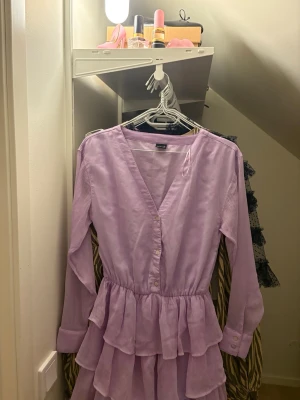 Ljuslila volangklänning med knappar - Supersöt ljuslila klänning från ginatricot med v-ringning, långa ärmar och volanger i flera lager. Klänningen har knappar framtill och på ärmsluten, samt ett lätt rynkat midjeparti. Materialet är tunt och har en lätt struktur som ger en luftig känsla.