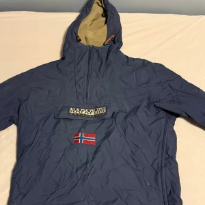 Vindjacka från Napapijri - Säljer en blå vindjacka från Napapijri med huva och halv dragkedja. Jackan har en stor ficka framtill med Napapijri-logga och ett broderat norskt flaggmärke. Insidan av huvan är beige och jackan har långa ärmar med resår vid ärmslut.